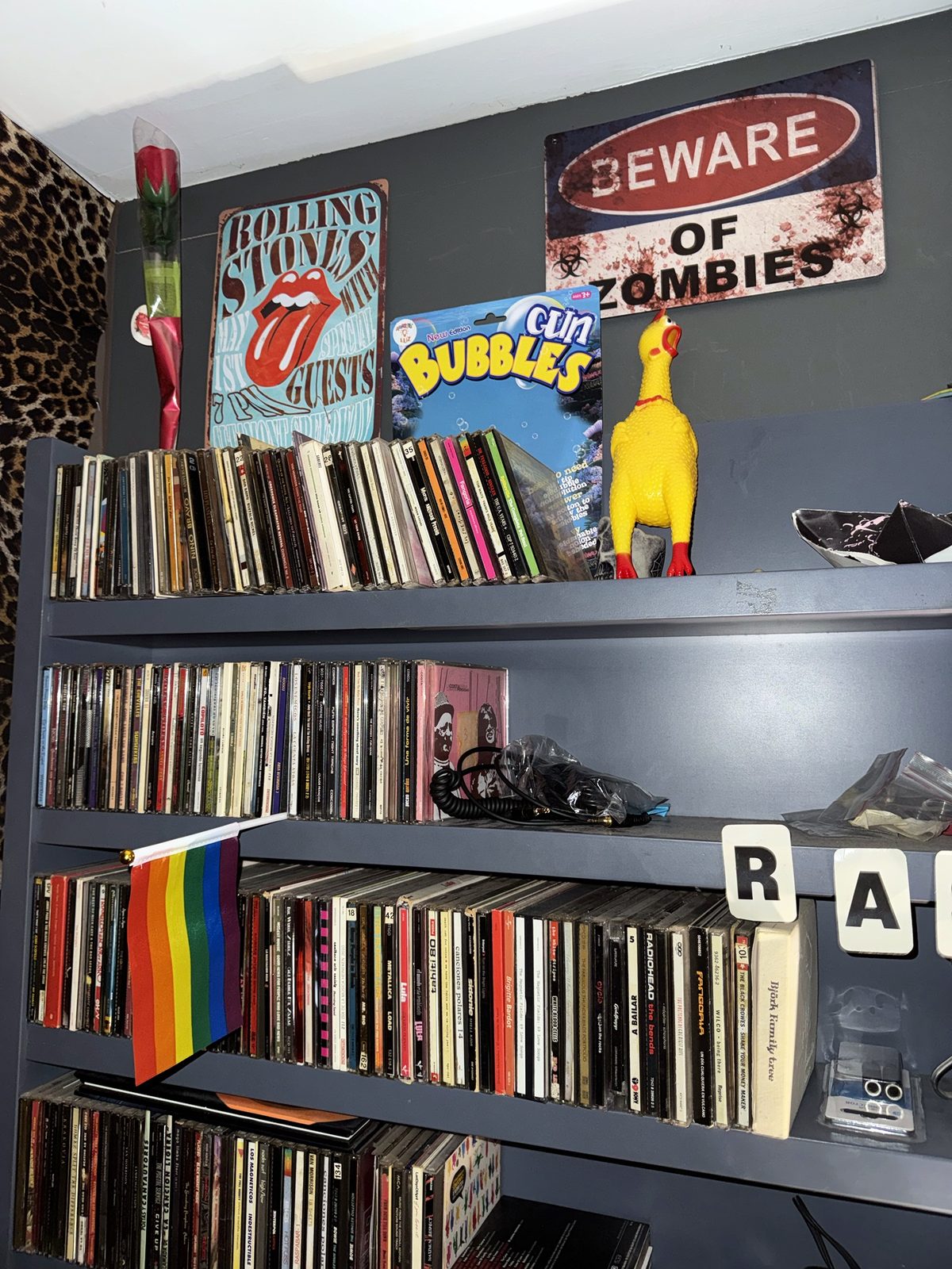 Estantería con CDs, cartel de los Rolling Stones, cartel de zombis y un pollo de goma