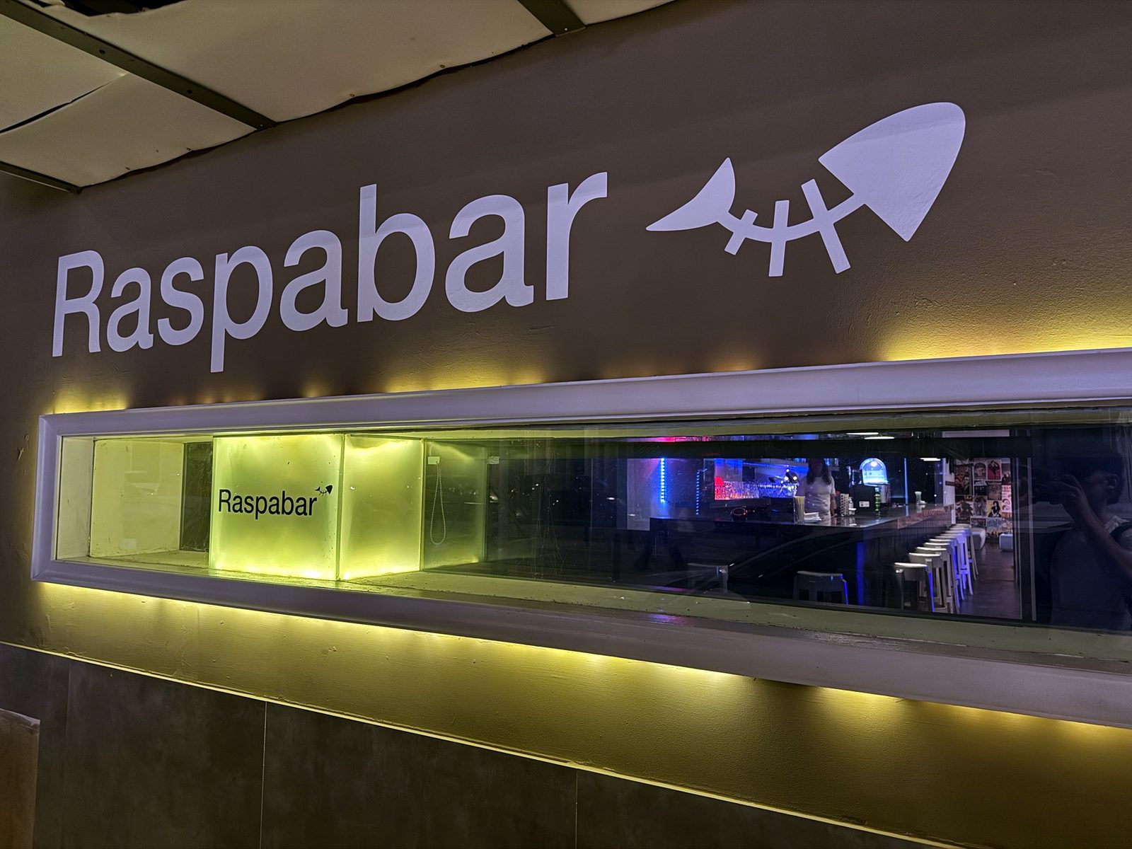 Letrero del Raspabar en la fachada, con la raspa de pescado y el escaparate iluminado